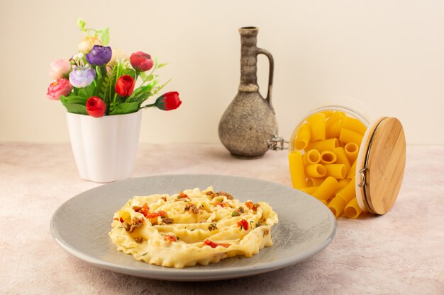 Vista frontal de una sabrosa comida de pasta italiana con verduras cocidas y pequeñas rebanadas de carne dentro de la placa gris junto con flores y pasta cruda en rosa