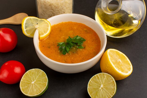 Vista frontal sabrosa sopa de verduras dentro de la placa con aceite de rodajas de limón y tomates rojos en el escritorio oscuro.