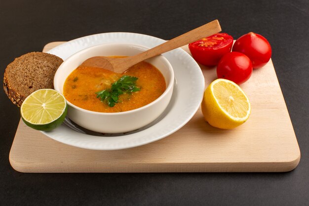 Vista frontal sabrosa sopa de verduras dentro de la placa con pan de pan, tomates de limón en el escritorio oscuro.