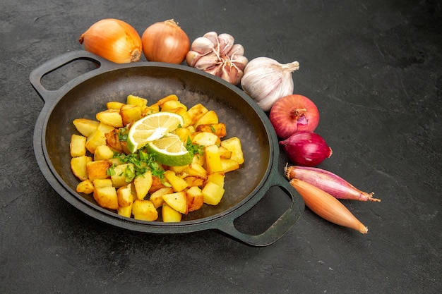 Vista frontal sabrosas patatas fritas dentro de la sartén con limón y ajos alrededor de la mesa oscura