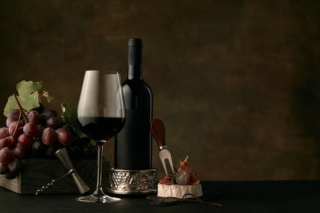 Vista frontal del sabroso plato de frutas de uvas con la botella de vino, queso y copa de vino en la oscuridad