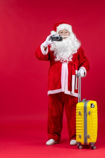 Vista frontal de santa claus con bolsa amarilla sosteniendo la cámara en la pared roja