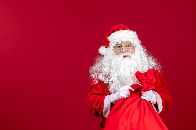 Vista frontal de santa claus con bolsa roja llena de regalos en el escritorio rojo emoción de navidad de vacaciones de año nuevo