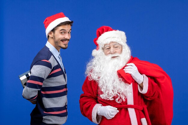 Vista frontal de santa claus con bolsa de transporte masculina joven llena de regalos en azul presente colores navideños navideños