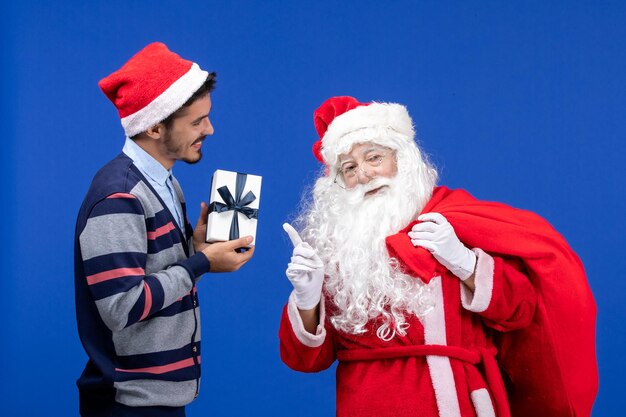 Vista frontal de santa claus con bolsa de transporte masculina joven llena de regalos en el piso azul presente de vacaciones