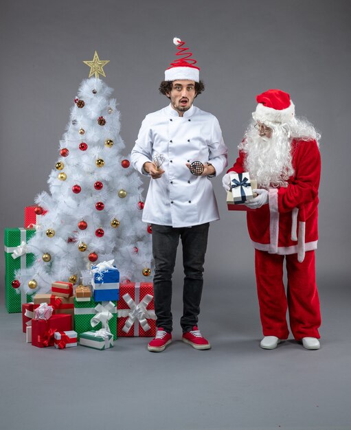 Vista frontal de santa claus con cocinero masculino alrededor de regalos de navidad en la pared gris