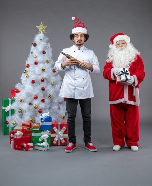 Vista frontal de santa claus con cocinero masculino alrededor de regalos de navidad en la pared gris