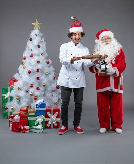 Vista frontal de santa claus con cocinero masculino alrededor de regalos de navidad en la pared gris