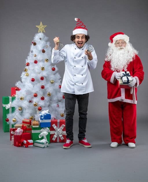 Vista frontal de santa claus con cocinero masculino alrededor de regalos de navidad en la pared gris