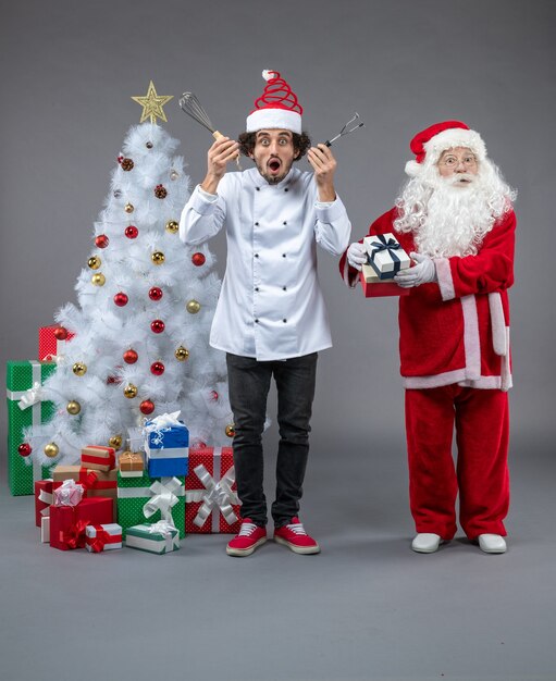 Vista frontal de santa claus con cocinero masculino alrededor de regalos de navidad en la pared gris