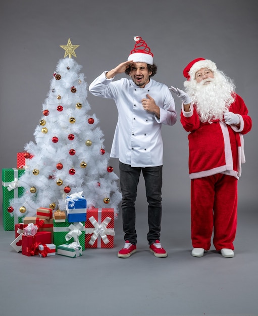 Vista frontal de santa claus con cocinero masculino alrededor de regalos de navidad en la pared gris