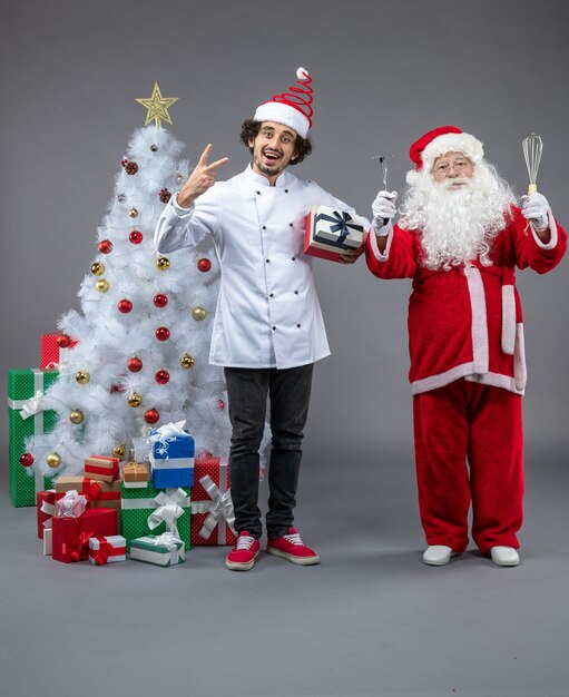 Vista frontal de santa claus con cocinero y regalos de navidad alrededor de la pared gris