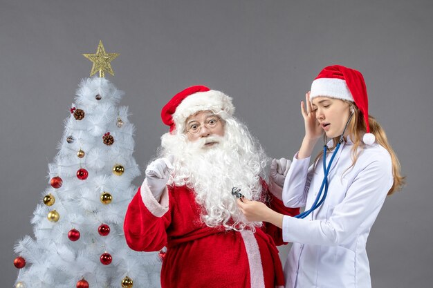 Vista frontal de santa claus con doctora que está comprobando su estado de salud en la pared gris