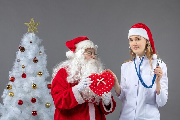 Vista frontal de santa claus con doctora que está observando presente con estetoscopio en la pared gris