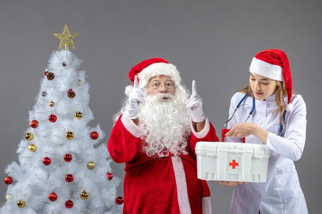 Vista frontal de santa claus con doctora que sostiene botiquín de primeros auxilios en la pared gris