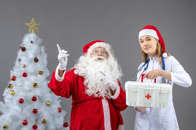 Vista frontal de santa claus con doctora que sostiene botiquín de primeros auxilios en la pared gris