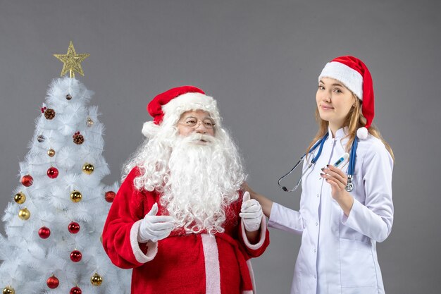 Vista frontal de santa claus con doctora que sostiene el dispositivo de medición de temperatura en la pared gris