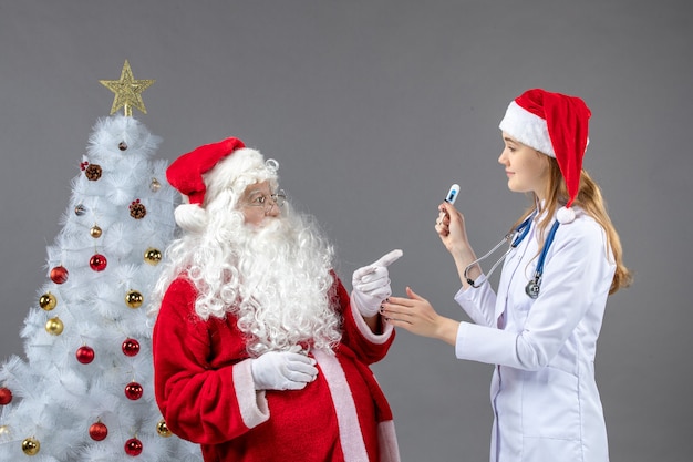 Vista frontal de santa claus con doctora que sostiene el dispositivo de medición de temperatura en la pared gris