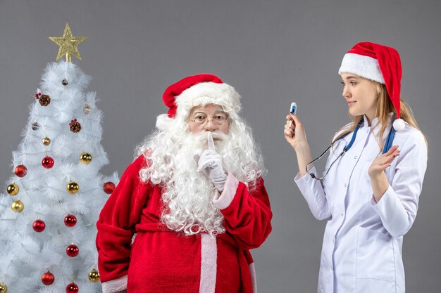 Vista frontal de santa claus con doctora que sostiene el dispositivo de medición de temperatura en la pared gris