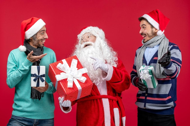 Vista frontal de santa claus con dos machos sosteniendo regalos en el escritorio rojo regalo de año nuevo emoción navidad rojo