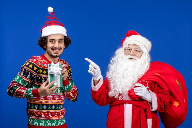 Vista frontal de santa claus con hombre joven y llevando una bolsa de regalos en color azul de vacaciones de navidad emoción