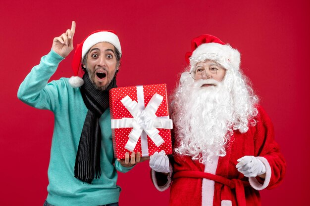 Foto gratuita vista frontal de santa claus con hombre sosteniendo presente de vacaciones en escritorio rojo emoción regalo rojo navidad