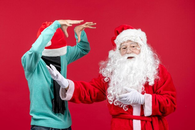 Vista frontal de santa claus con joven asustándolo en la pared roja