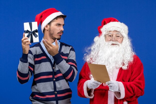 Vista frontal de santa claus con joven leyendo carta y sosteniendo presente en la pared azul