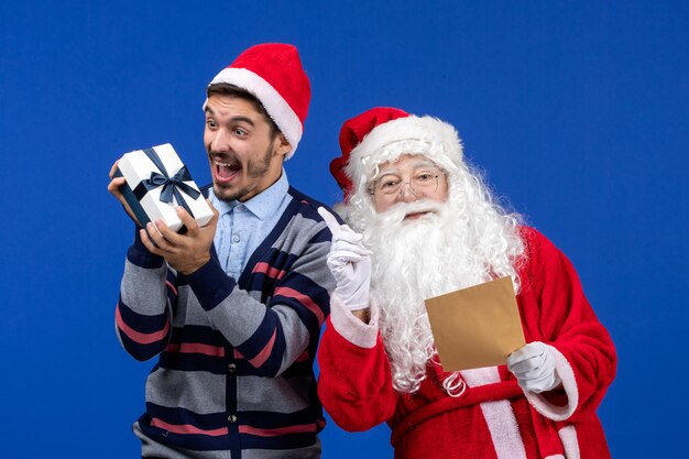 Vista frontal de santa claus con joven leyendo carta y sosteniendo presente en la pared azul