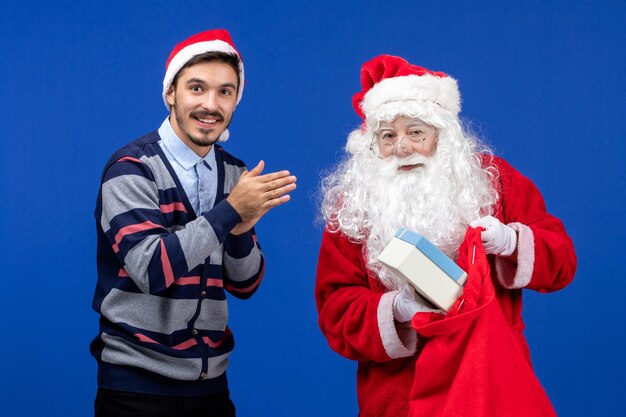 Vista frontal de santa claus con joven sosteniendo la bolsa de regalos en la pared azul