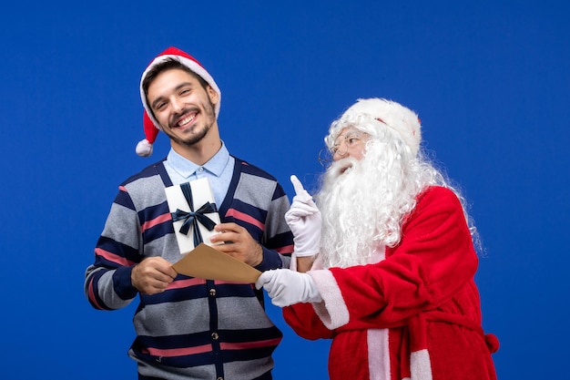 Foto gratuita vista frontal de santa claus con joven sosteniendo carta y presente en la pared azul