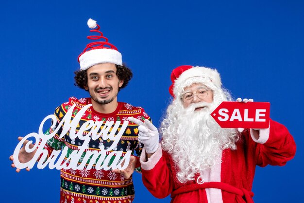 Vista frontal de santa claus con joven sosteniendo feliz navidad y escritos de venta en la pared azul