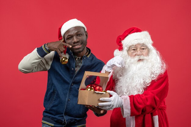 Vista frontal de santa claus con joven sosteniendo juguetes de árbol de Navidad en pared roja