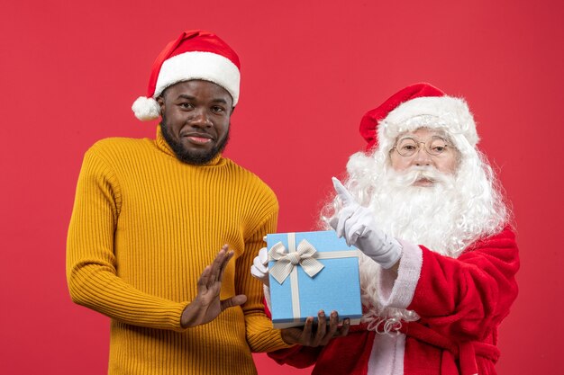 Vista frontal de santa claus con joven sosteniendo presente en la pared roja