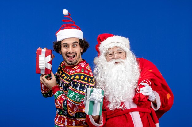 Vista frontal de santa claus con joven sosteniendo regalos de navidad en la pared azul