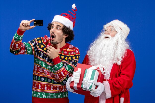 Vista frontal de santa claus con joven sosteniendo regalos y tarjeta bancaria en la pared azul