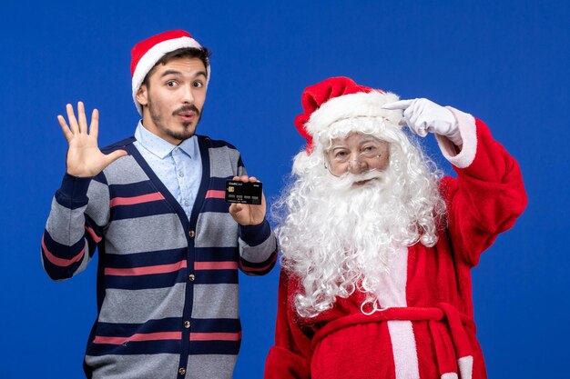 Vista frontal de santa claus con joven sosteniendo una tarjeta bancaria en la pared azul