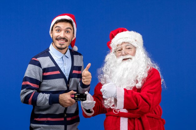 Vista frontal de santa claus con joven sosteniendo una tarjeta bancaria en la pared azul