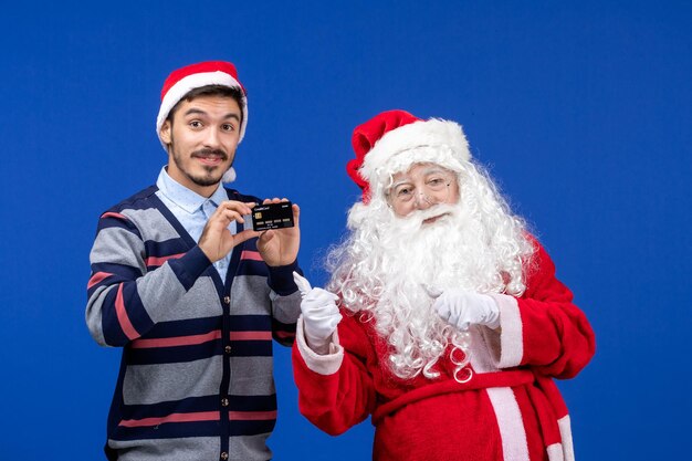 Vista frontal de santa claus con joven sosteniendo una tarjeta bancaria en la pared azul