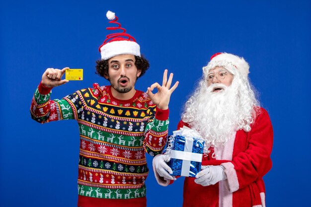 Vista frontal de santa claus con joven sosteniendo tarjeta bancaria y presente en la pared azul