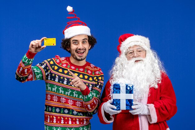 Vista frontal de santa claus con joven sosteniendo tarjeta bancaria y presente en la pared azul