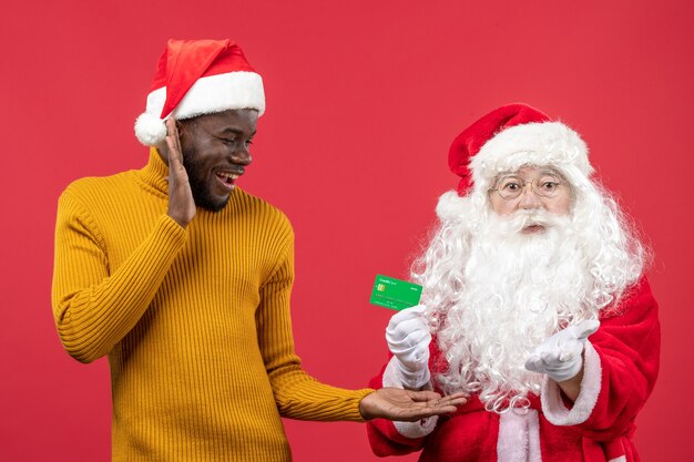 Vista frontal de santa claus con joven sosteniendo tarjeta bancaria verde en la pared roja