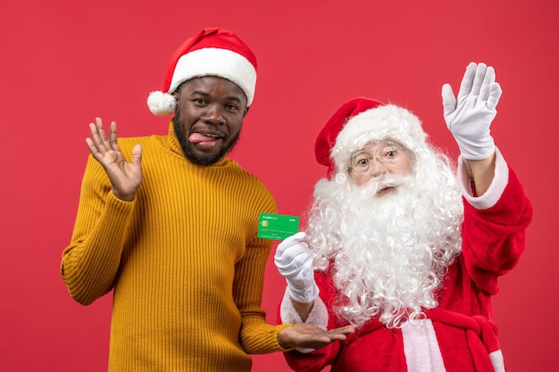 Vista frontal de santa claus con joven sosteniendo tarjeta bancaria verde en la pared roja