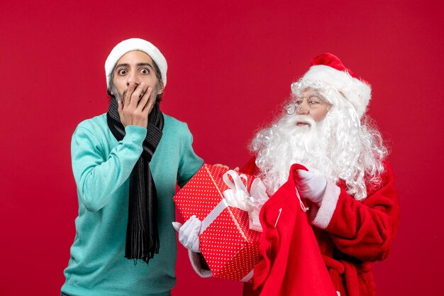 Vista frontal de santa claus con macho sacando el presente de la bolsa en el color rojo de vacaciones de emoción de Navidad