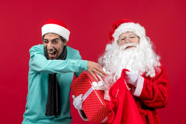 Foto gratuita vista frontal de santa claus con macho sacando el presente de la bolsa en el escritorio rojo color de vacaciones de emoción de navidad