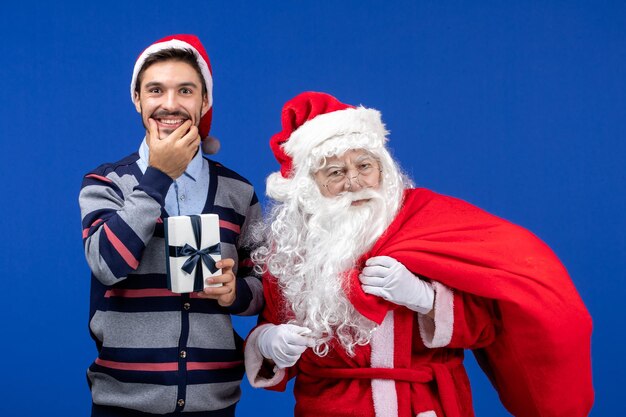 Vista frontal de santa claus con machos jóvenes bolsa llena de regalos en color azul vacaciones presenta navidad