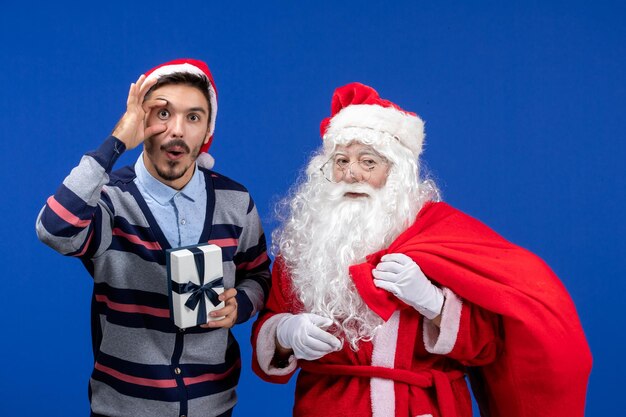 Vista frontal de santa claus con machos jóvenes bolsa de transporte llena de regalos en color azul Navidad presente de vacaciones