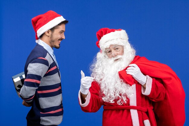 Vista frontal de santa claus con machos jóvenes bolsa de transporte llena de regalos en el escritorio azul color de navidad presente de vacaciones