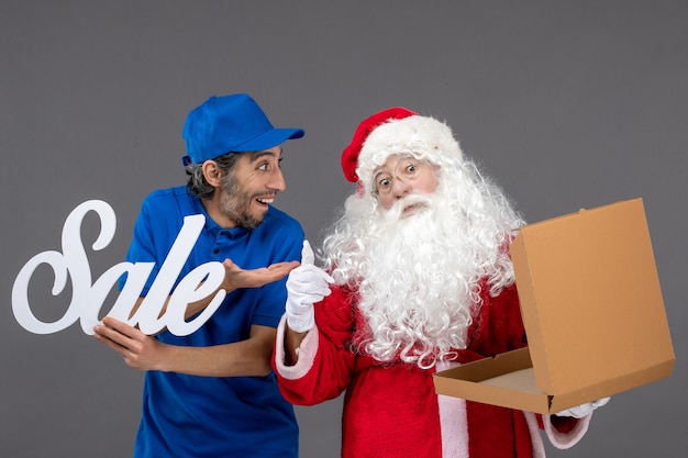 Vista frontal de santa claus con mensajero masculino con escritura de venta y caja de comida vacía en la pared gris