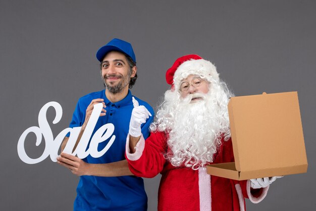 Vista frontal de santa claus con mensajero masculino con escritura de venta y caja de comida vacía en la pared gris